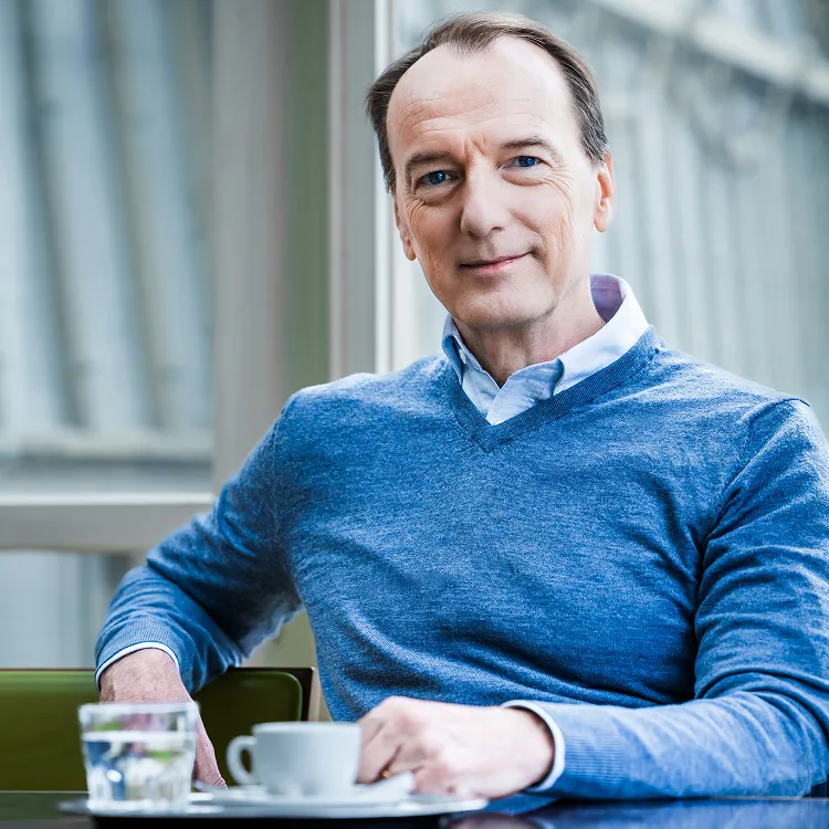 Portrait of Marc Elsberg in a blue sweater, smiling at a table with a coffee cup.