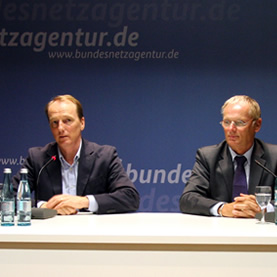 Marc Elsberg sitzt am Rednerpult vor einem Plakat mit der Aufschrift www.bundesnetzagentur.de