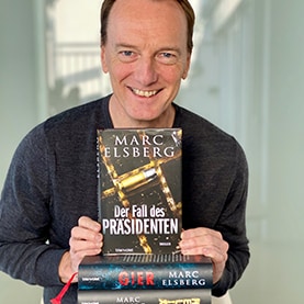 Marc Elsberg hält seinen Roman „Der Fall des Präsidenten“,lächelnd mit zwei Händen in die Kamera.