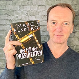 Marc Elsberg hält „Der Fall des Präsidenten“ in einer Hand und lächlt in die Kamera.