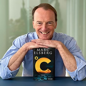 Marc Elsberg legt beide Hände und sein Kinn auf das Buch Celsius, während er in die Kamera blickt.