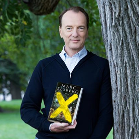 Marc Elsberg steht im Park an einen Baum gelehnt und hält sein Buch mit einem gelben X in der Hand.
